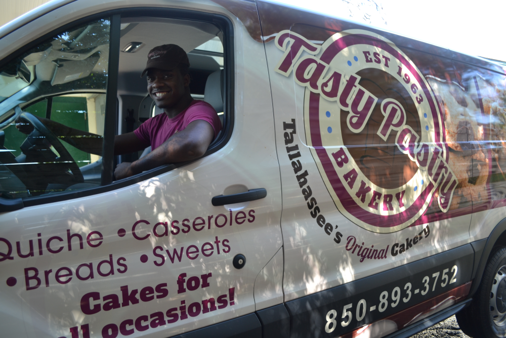 A man wearing Tasty Pastry's uniform smiles from the driver's seat of a Tasty Pastry delivery van